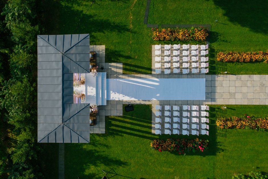 An aerial view of an outdoor wedding ceremony setup in a lush green garden. A white floral aisle runner extends from a shaded gazebo decorated with pink and white flowers, leading to rows of white cha