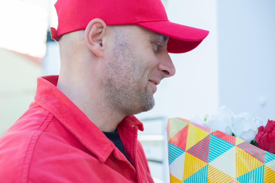 A man wearing a red cap and matching red jacket is holding a colorful, geometric-patterned gift bag filled with fresh flowers, including white blooms and rich pink roses, in an outdoor setting with so