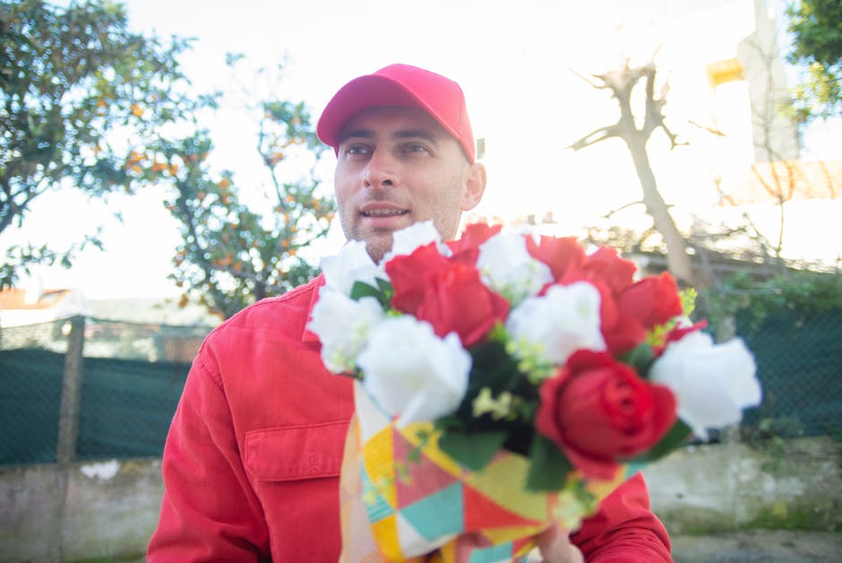 A male florist wearing a red baseball cap and matching red jacket is outdoors, holding a colorful bouquet of fresh flowers that include red and white roses with lush green leaves. The bouquet is wrapp