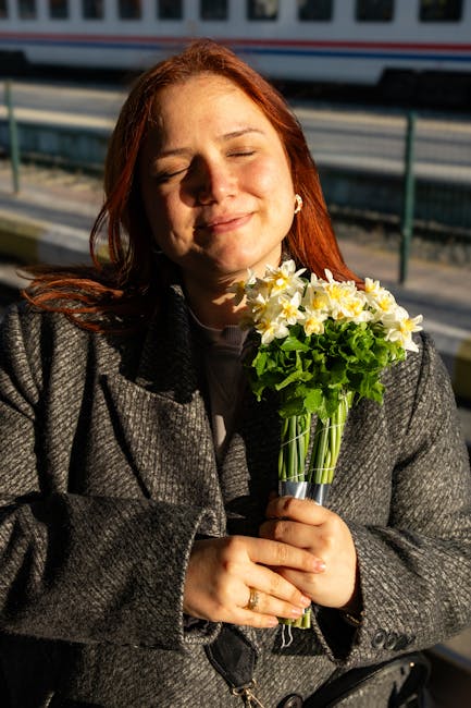 A woman with red hair and a content expression is holding a bouquet of fresh flowers, including white and yellow daffodils and green foliage, wrapped in clear plastic. She is outdoors, with sunlight i
